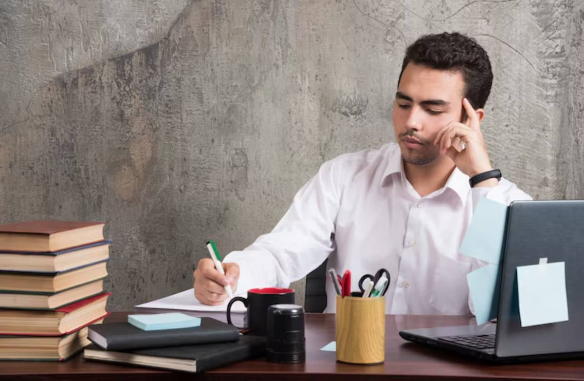 A person in a white shirt sits at a desk, jotting notes with books, a laptop, and office supplies surrounding them.