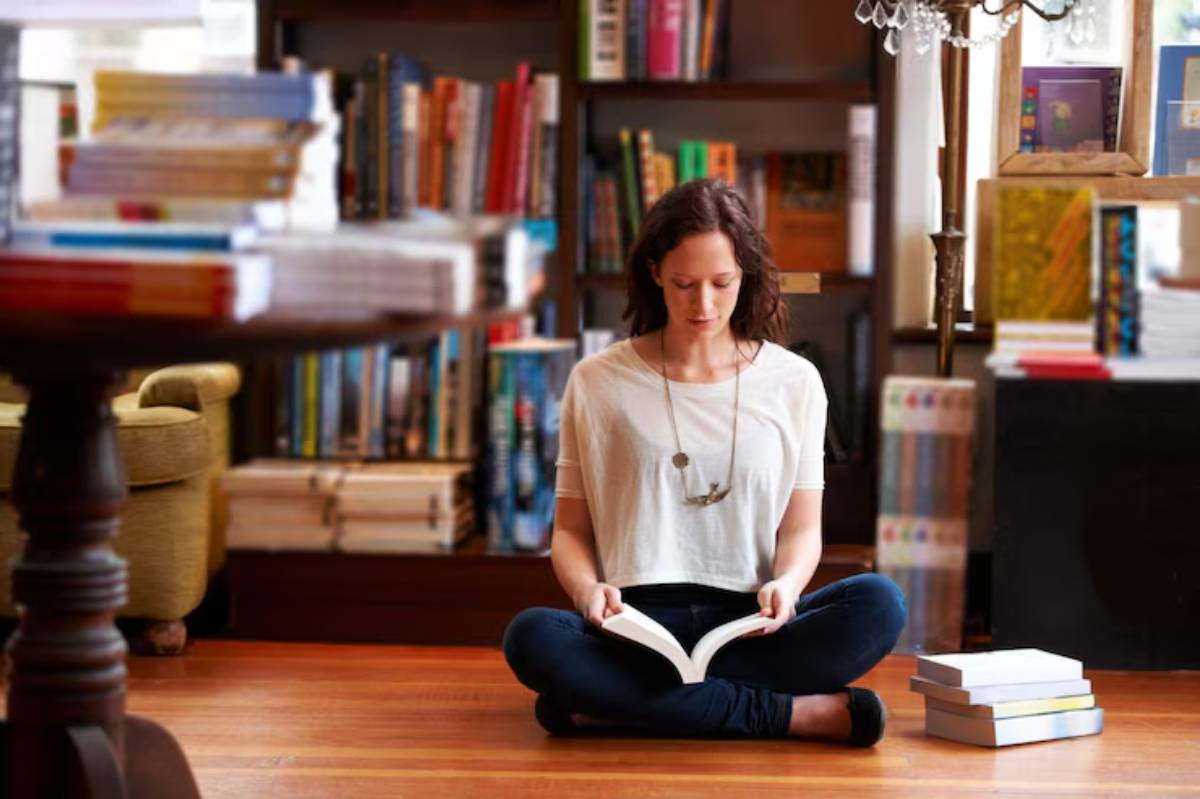 A person sitting cross-legged on the floor in a cozy library, reading a book, with shelves full of colorful books in the background.