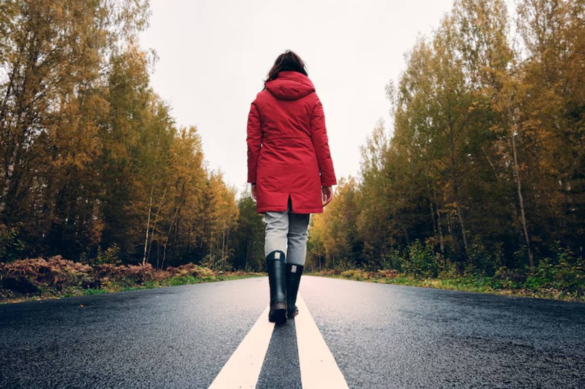 A woman in a red coat walks alone on a deserted road, surrounded by vibrant autumn trees under a cloudy sky.