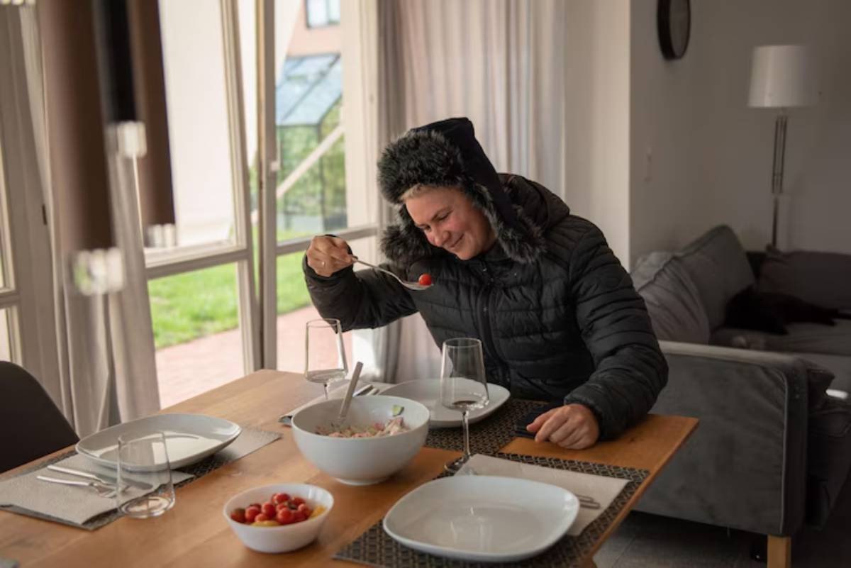 A person in a black puffer jacket sits at a dining table with plates, glasses, and bowls of food, preparing to eat a cherry tomato.
