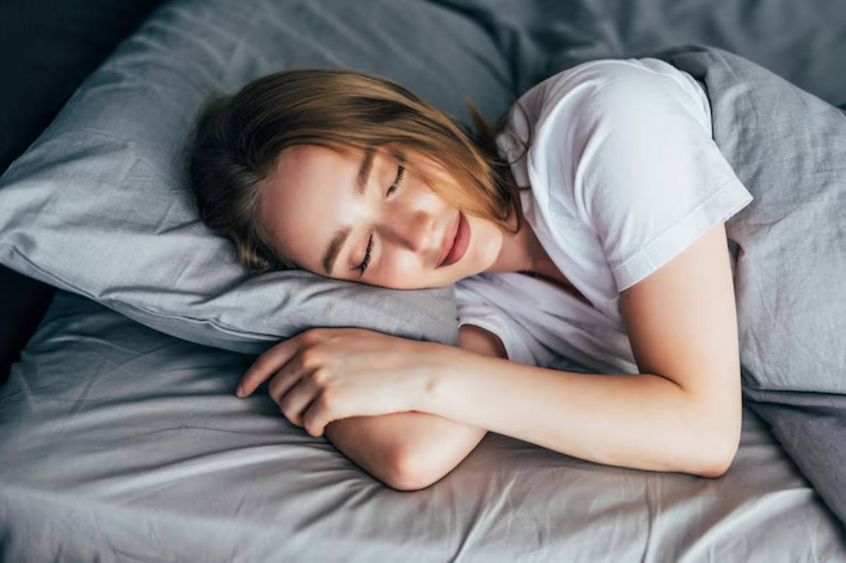 A person lies peacefully on a bed, resting their head on a plush pillow, with arms crossed comfortably. Soft gray bedding surrounds them.