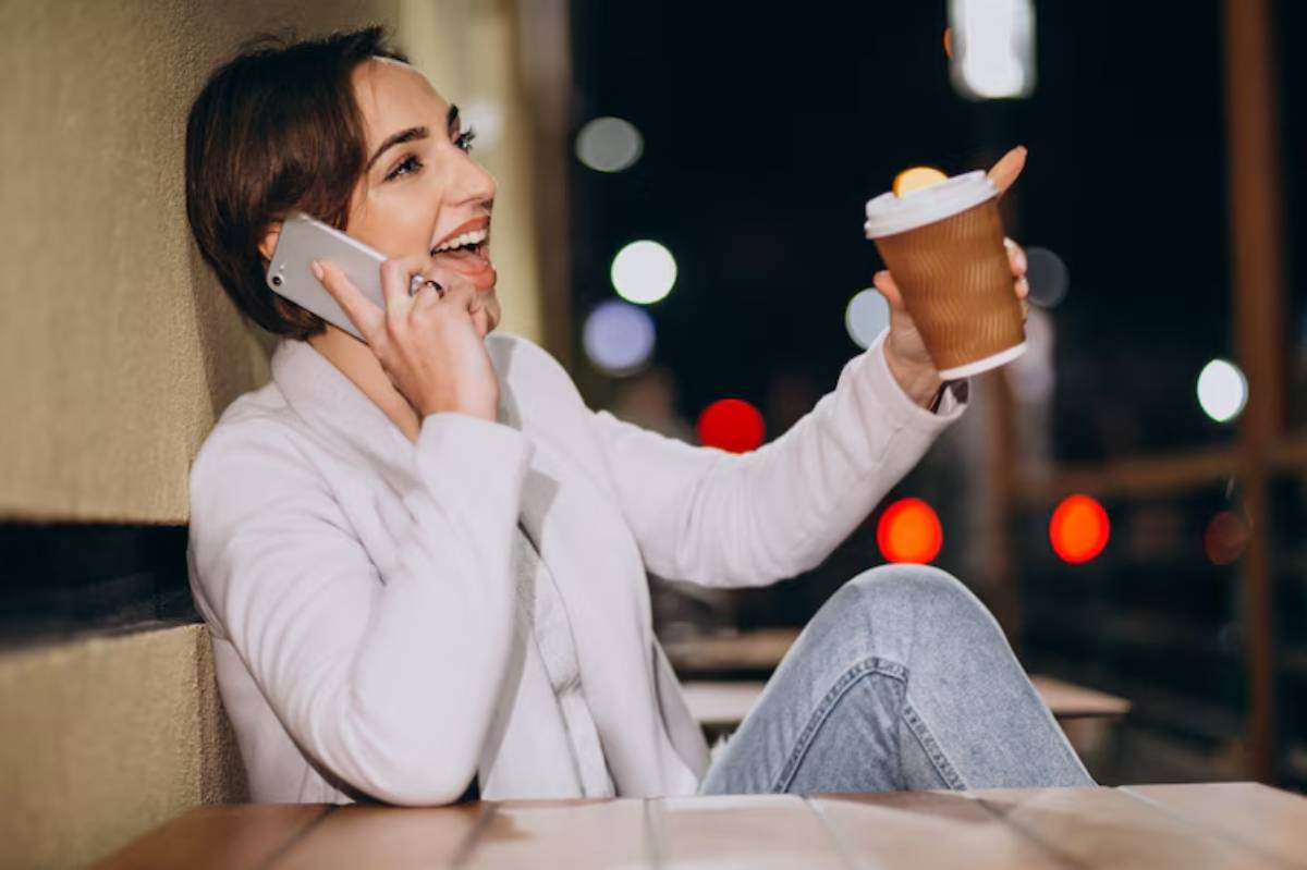 A person in a white jacket sits at a table, holding a coffee cup and talking on a phone in a softly lit outdoor space at night.