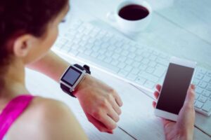 A person is sitting at a desk with a smartwatch on their wrist and holding a smartphone, alongside a cup of coffee and a keyboard.