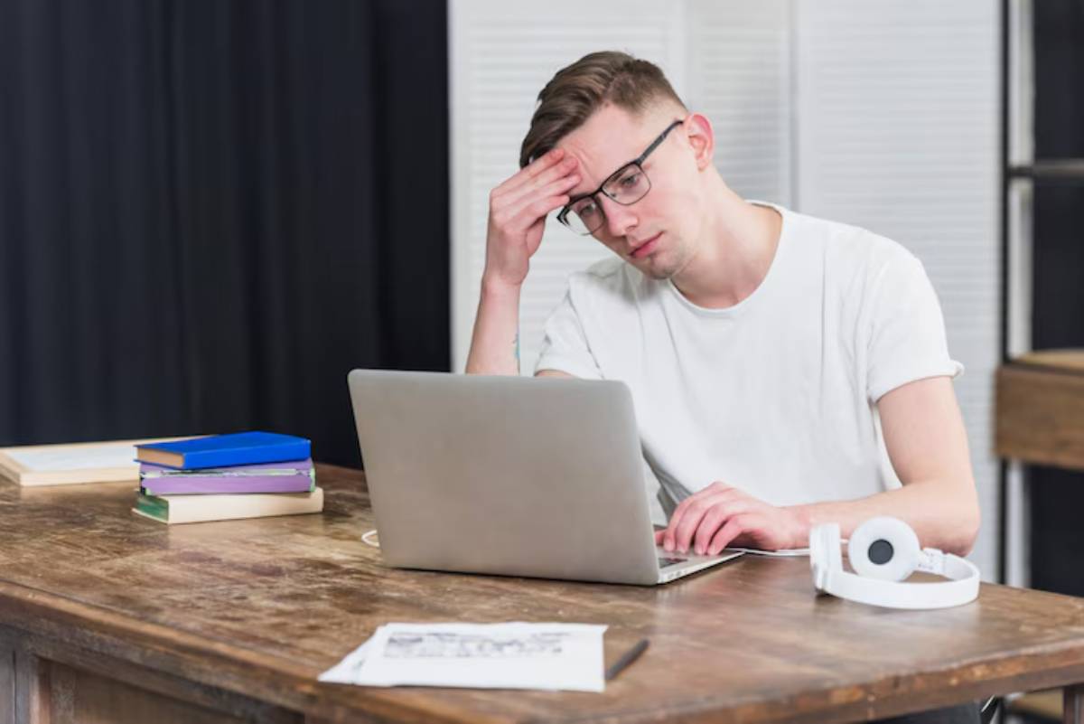 A young man sits at a wooden desk, looking stressed while working on a laptop, surrounded by books and headphones.