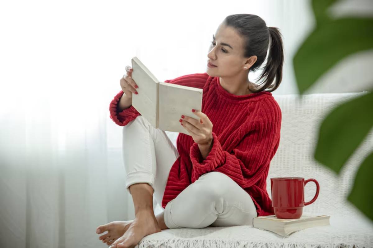A person sits comfortably on a couch, reading a book, dressed in a red sweater and white pants, with a red mug beside them.
