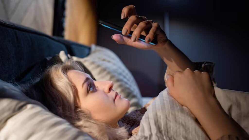 A person lounging in bed, holding a smartphone, illuminated by soft light, surrounded by cozy bedding.