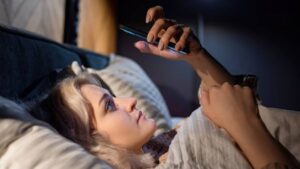 A person lounging in bed, holding a smartphone, illuminated by soft light, surrounded by cozy bedding.