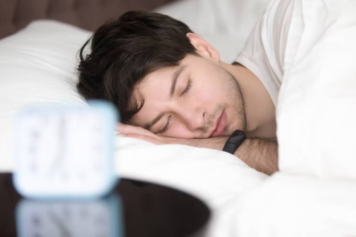 A person is peacefully sleeping on a bed, surrounded by fluffy white pillows, with a bedside clock visible in the foreground.