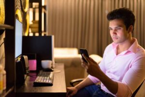 A young man in a pink shirt sits at a desk, holding a smartphone, with a computer and speakers visible in a cozy workspace setting.