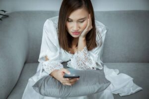 A person in a white lace robe sits on a gray sofa, holding a smartphone and resting on a gray pillow, looking contemplative.