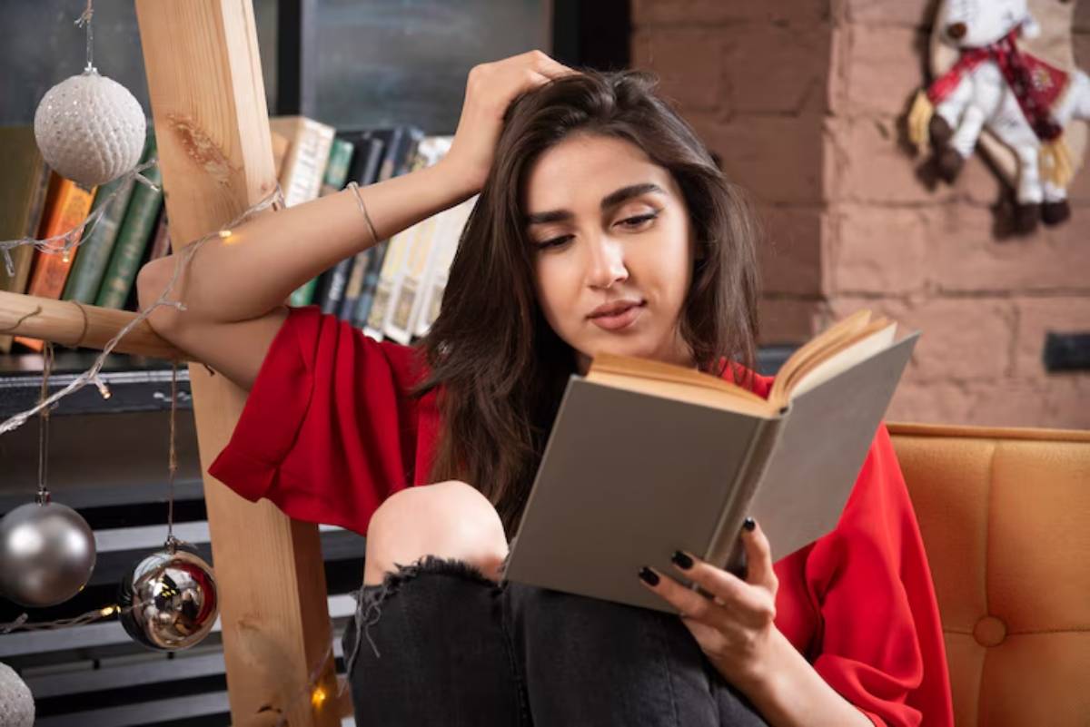 A young woman in a red shirt sits on a couch, absorbed in reading a book, surrounded by bookshelves and festive decor.