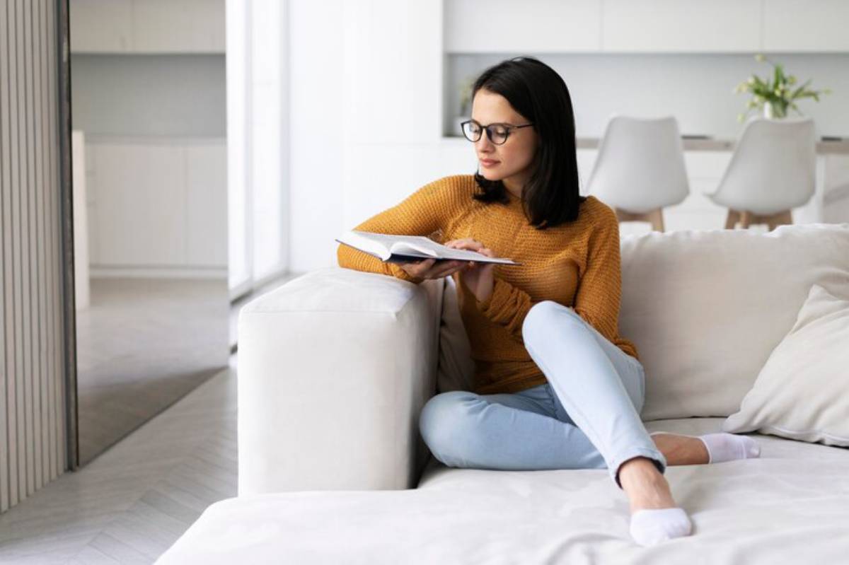 A person in a cozy, mustard sweater relaxes on a white couch, reading a book in a light-filled, modern living space.