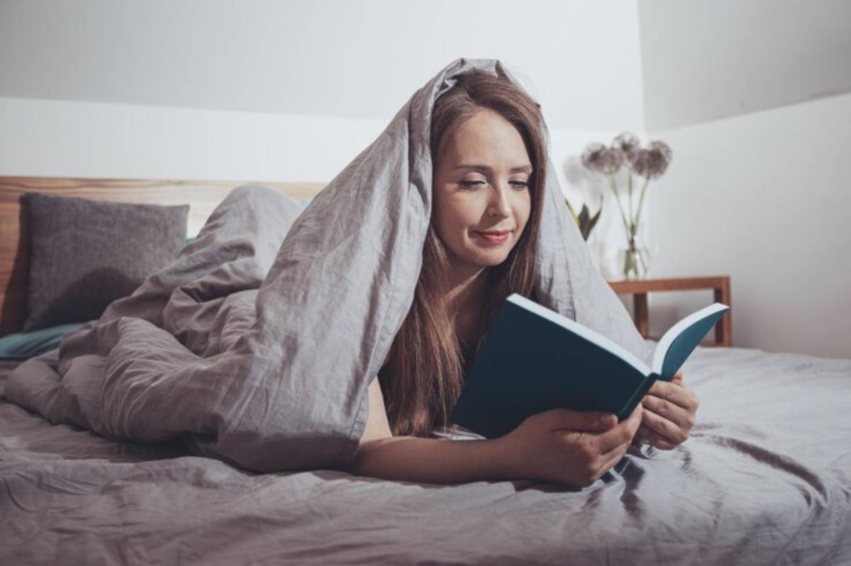 A person wrapped in a gray blanket lies on a bed, reading a blue book in a cozy, softly lit room with decorative flowers nearby.
