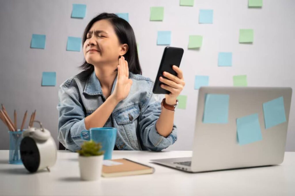 A person in a denim jacket holds a smartphone, seated at a desk with a laptop, mug, notebook, and potted plant, surrounded by sticky notes.