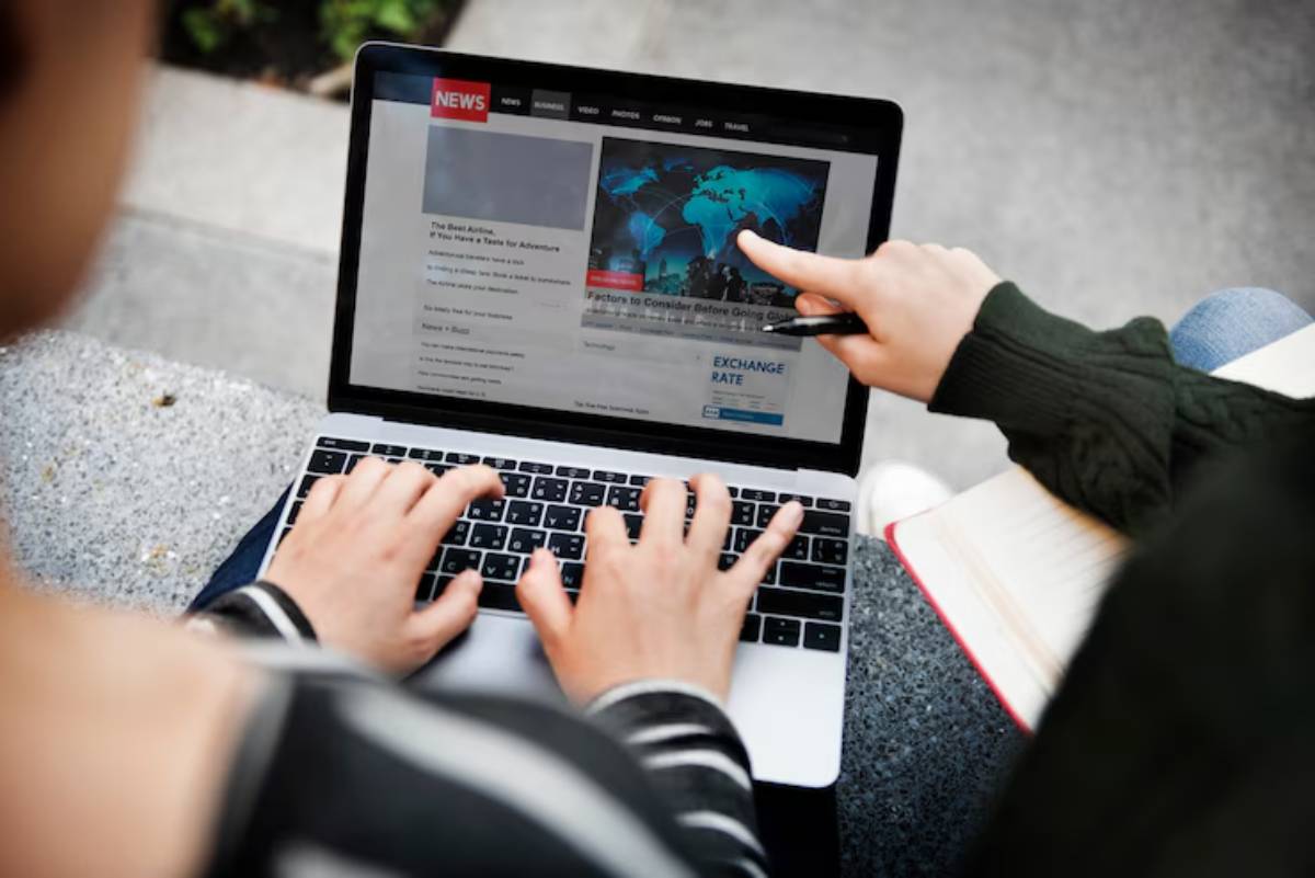 Two people engage in a discussion over a laptop displaying news articles, one pointing to a map of the world on the screen.